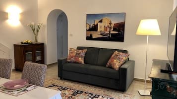 Un soggiorno con un divano scuro, cuscini decorati, un quadro raffigurante un edificio, una lampada e una zona pranzo con tavolo e sedie.