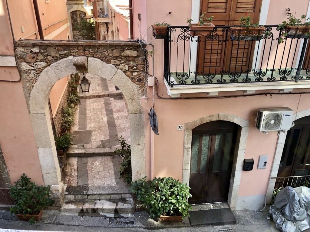 Un arco in pietra conduce all'ingresso di un edificio, con una porta nera e un piccolo balcone adornato con vasi di piante.