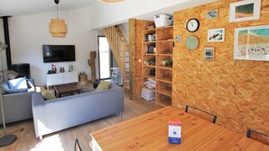 A modern living room with a sofa, a dining table, and a TV. There is a bookshelf and a staircase leading to another level. - Villa Les Hibiscus - 500m from the beach - authentic style and design for 8 (Longeville-sur-Mer)