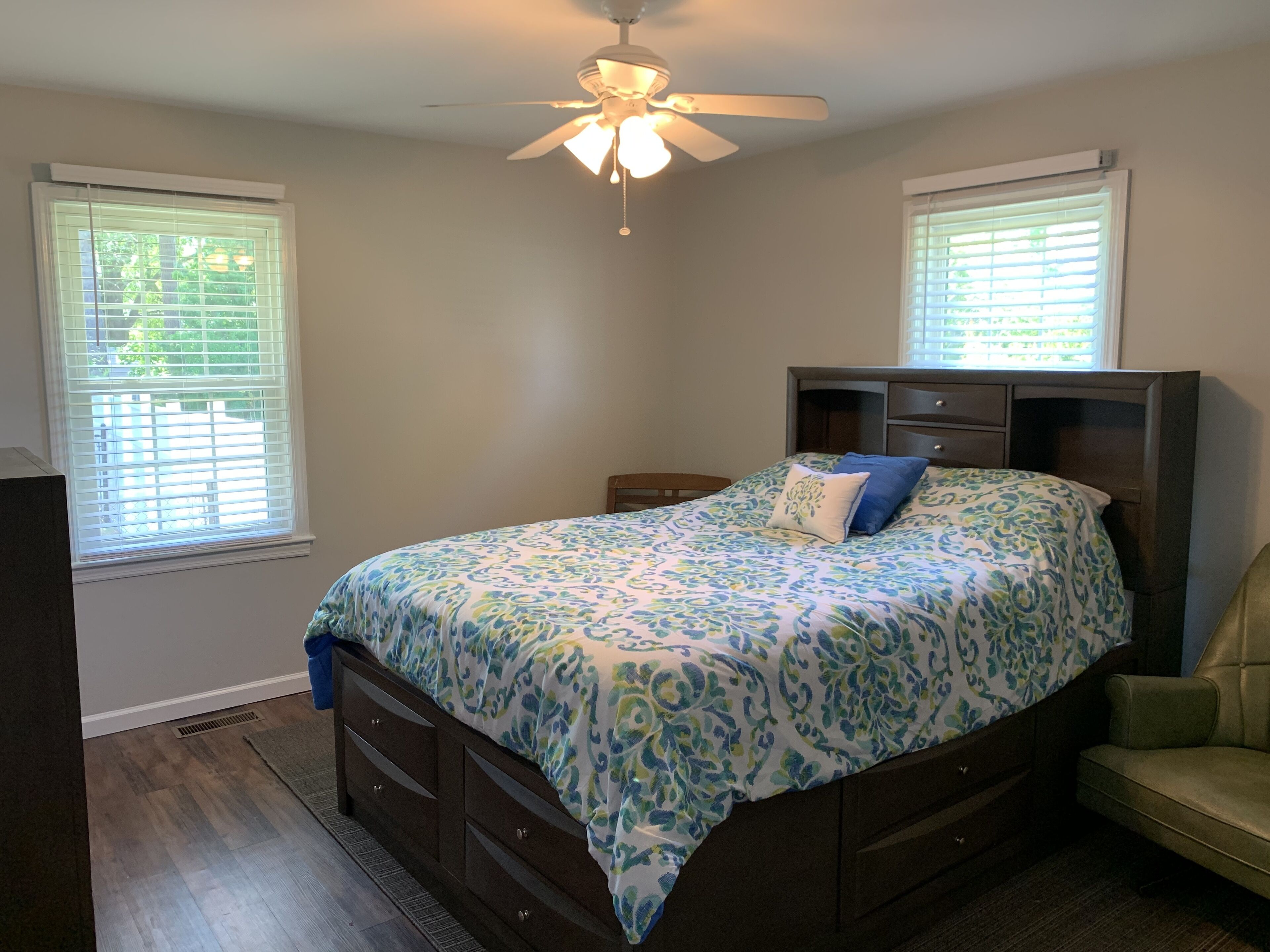 A bedroom with a bed, two windows with blinds, a ceiling fan, and a nightstand.
