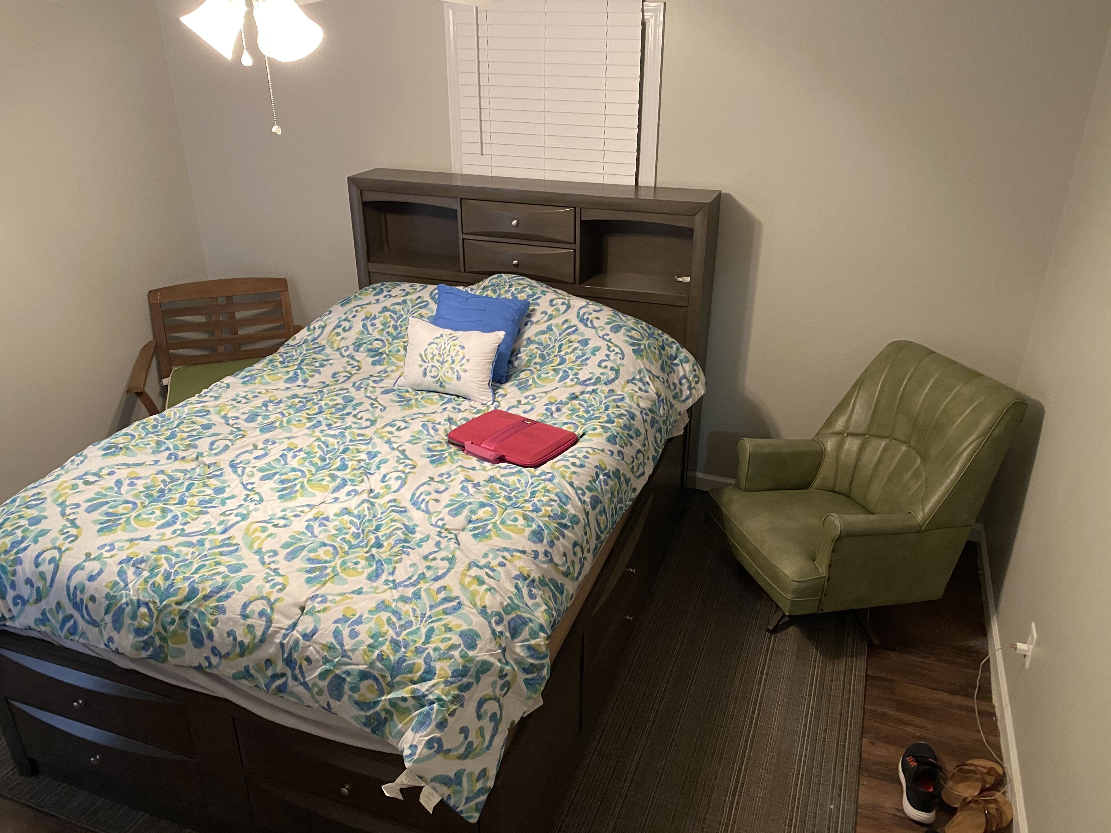 A bedroom with a bed, a green chair, a desk, and a window with blinds.