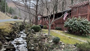 Una cabana rústica amb un balcó de fusta, un petit torrent que flueix per un llit rocos i un paisatge forestal de fons.