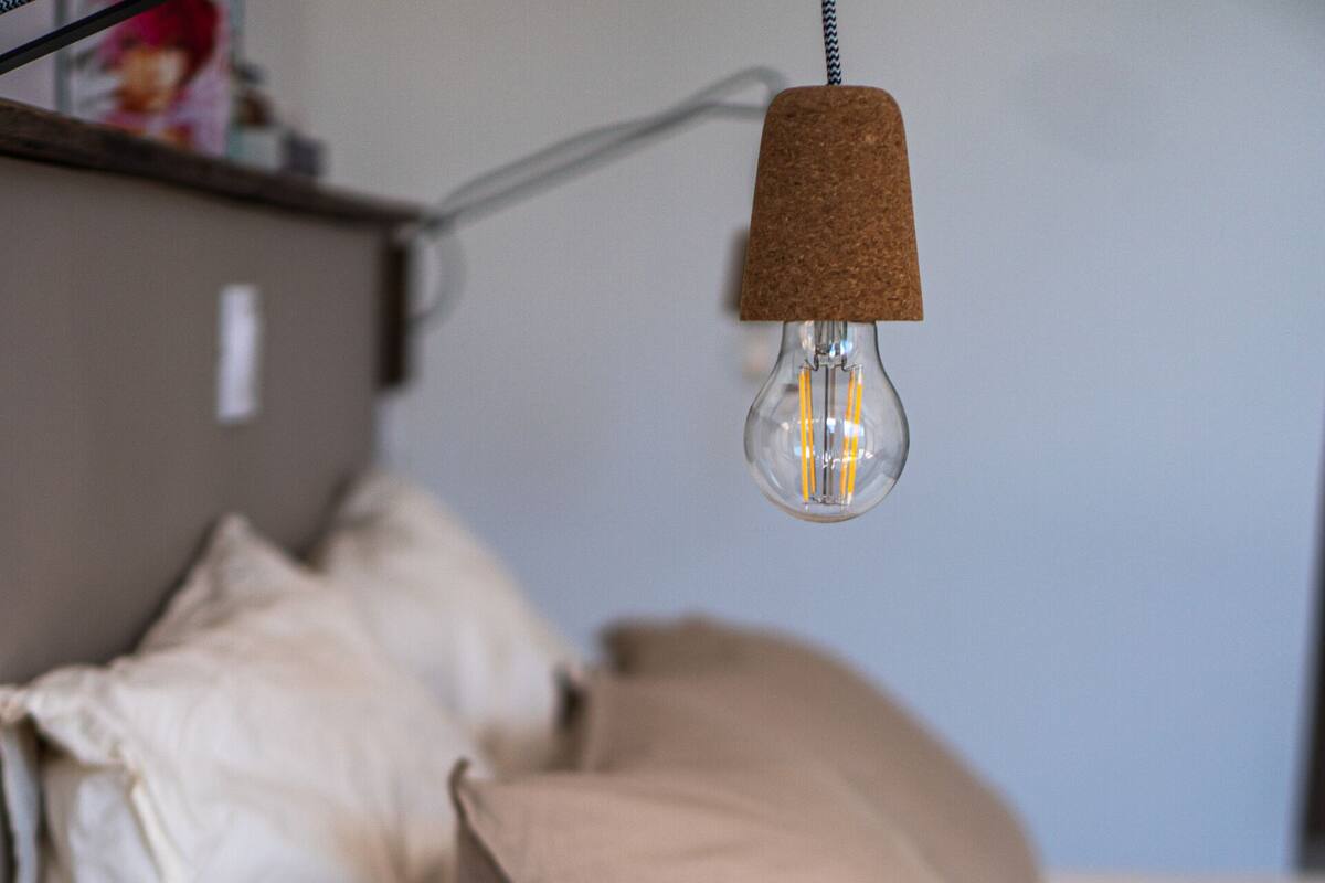 A bed with white and beige bedding, a cork lamp shade, and a visible power cord.