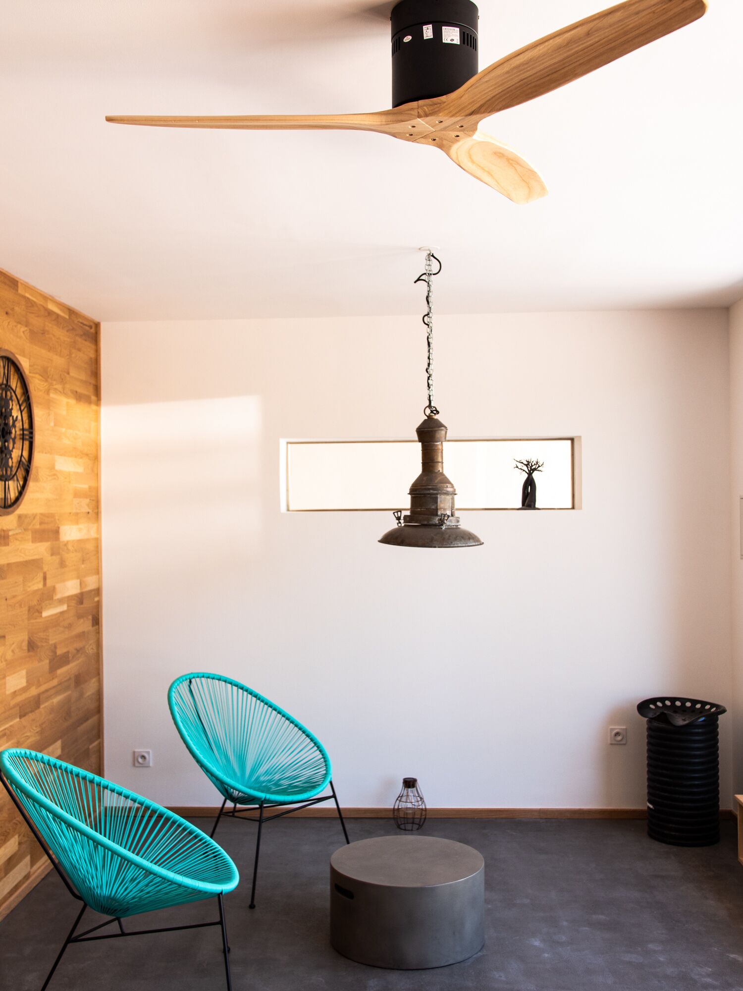 A modern room with a ceiling fan, two turquoise chairs, a round ottoman, a hanging light fixture, and a framed artwork on the wall.