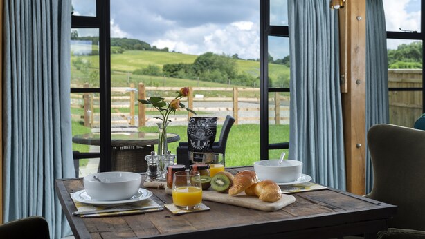 Una mesa de desayuno con un bol, croissants, jugo y vistas a un paisaje herboso a través de amplios ventanales.