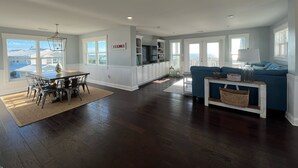 A spacious living area with a dining table, blue sofa, and a kitchen with white cabinets. - Ocean View - Family Friendly - Steps to Beach (Bald Head Island)
