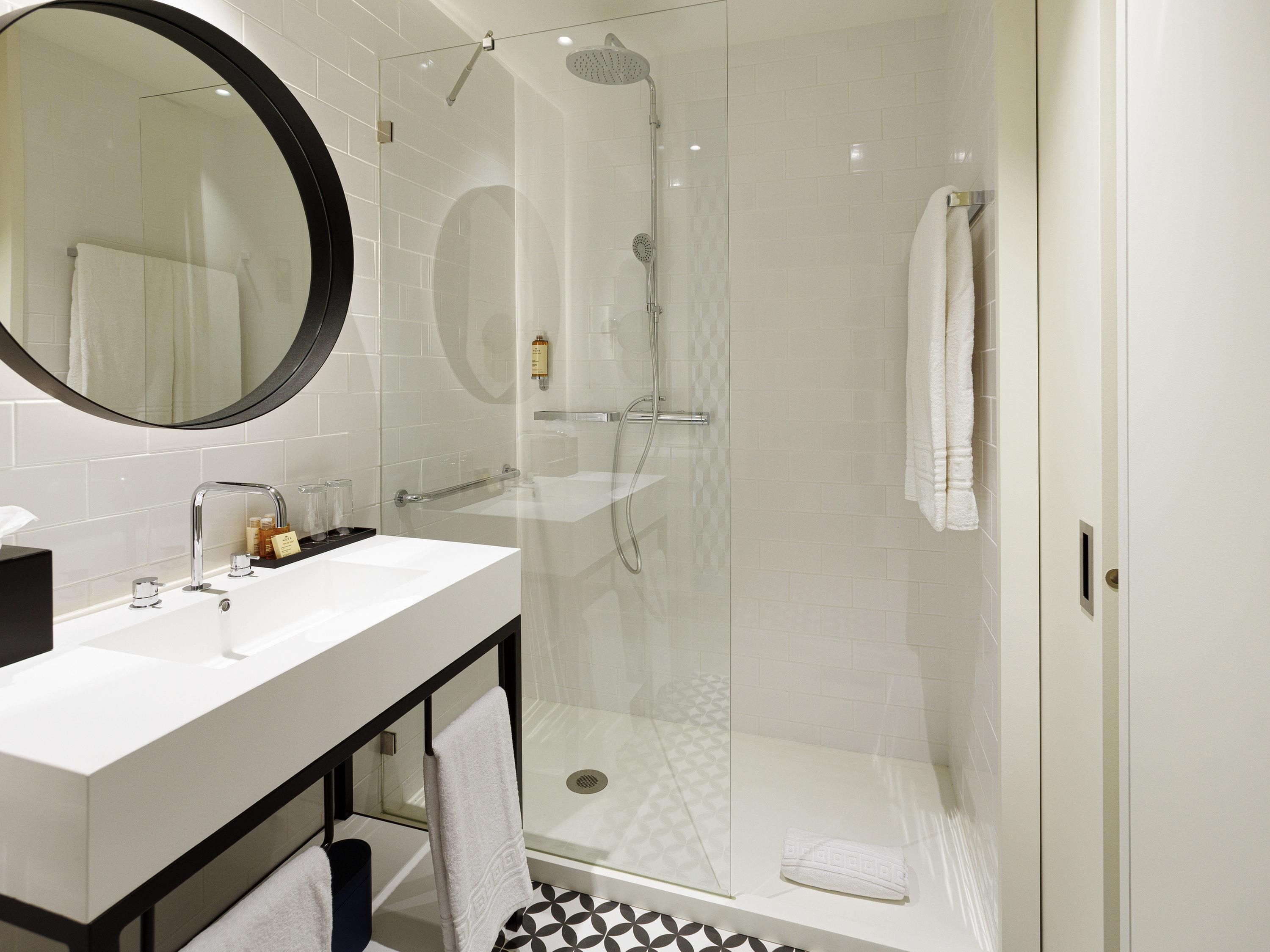 Une salle de bain moderne avec une douche à l’italienne en verre, un lavabo blanc doté d’un plan de travail noir et un miroir rond placé au-dessus du lavabo.