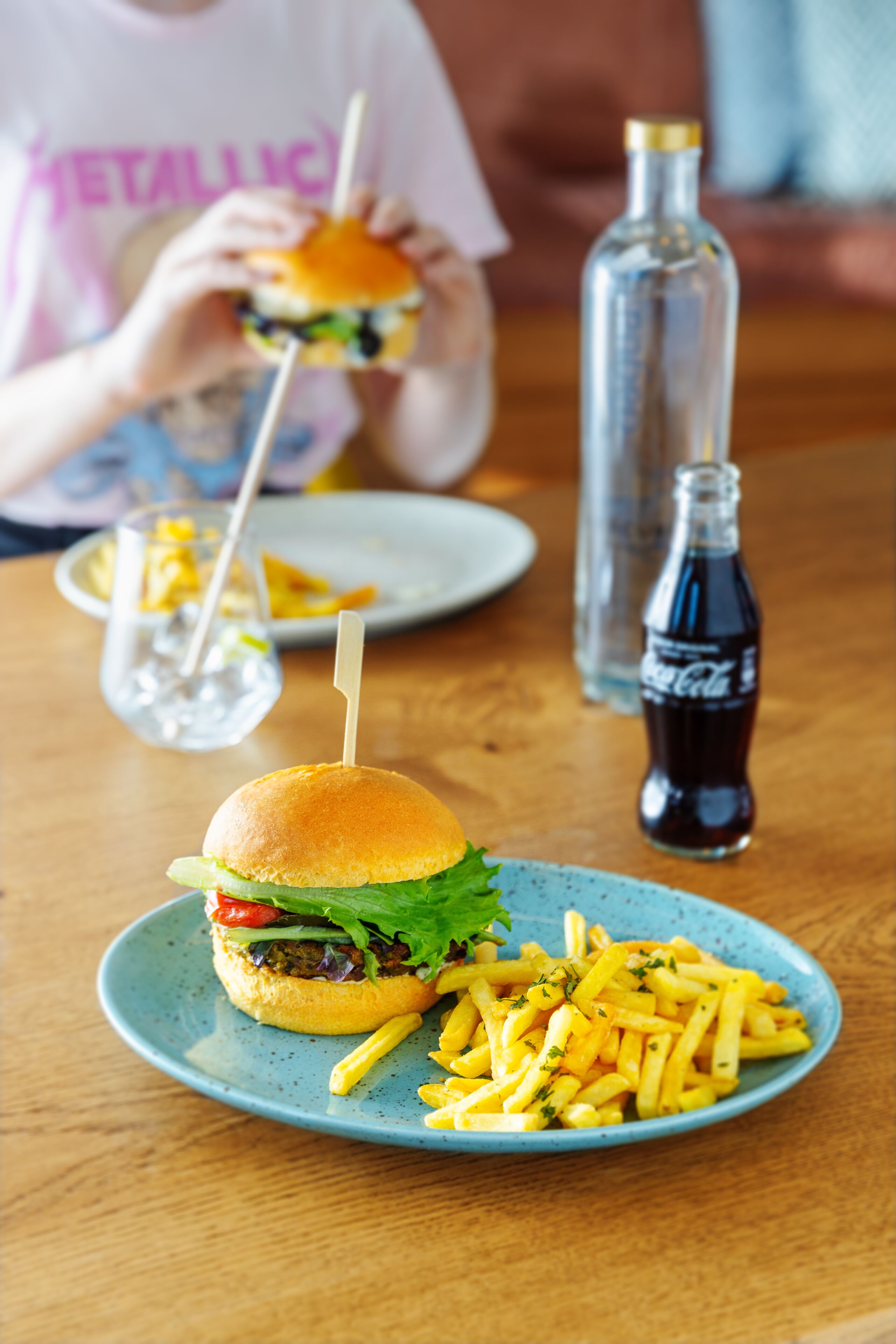 A meal with a burger and fries on a plate, a bottle of soda, and a glass of water on a table.