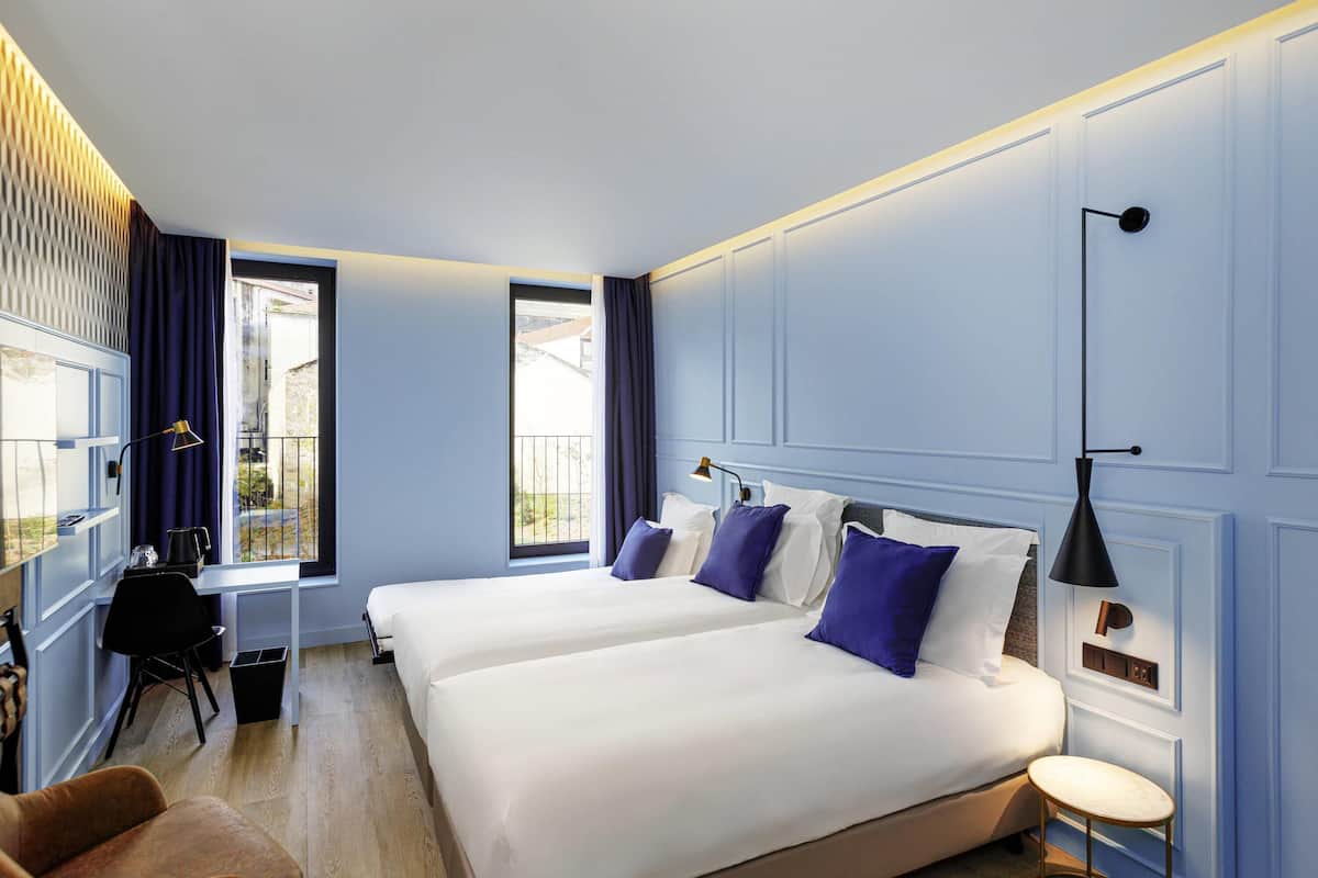 A hotel room with two beds, a desk, a chair, a small table, and a view of the outdoors.