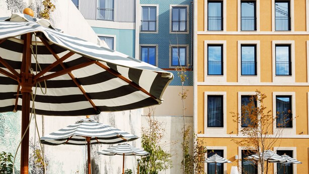 A row of colorful buildings with balconies, some with plants. There are striped umbrellas in the foreground.