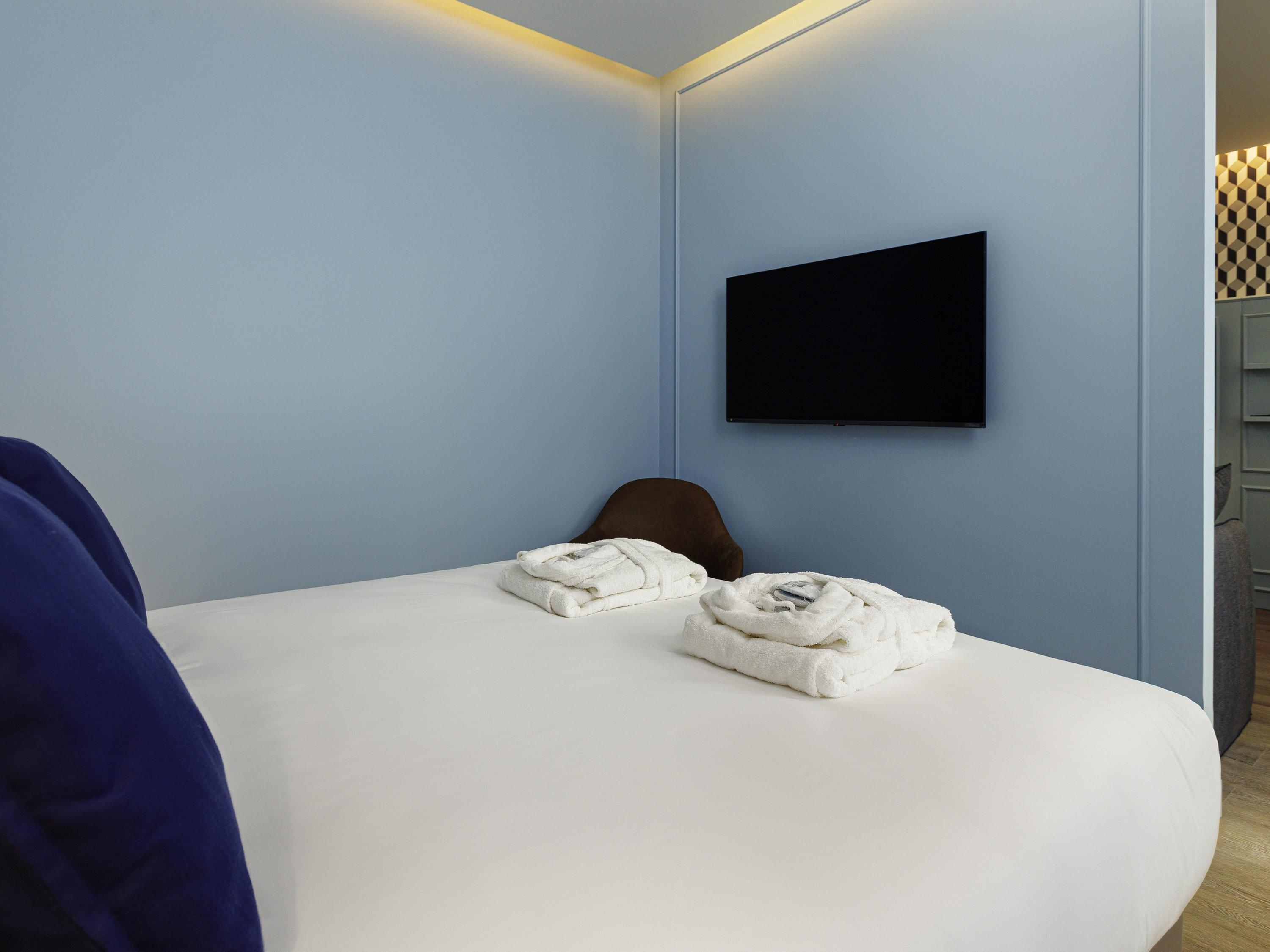 A hotel room with a bed, a flat-screen TV mounted on the wall, and a chair.