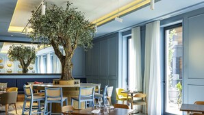 A restaurant interior with a large tree centerpiece, blue and yellow chairs, and a geometric patterned floor.