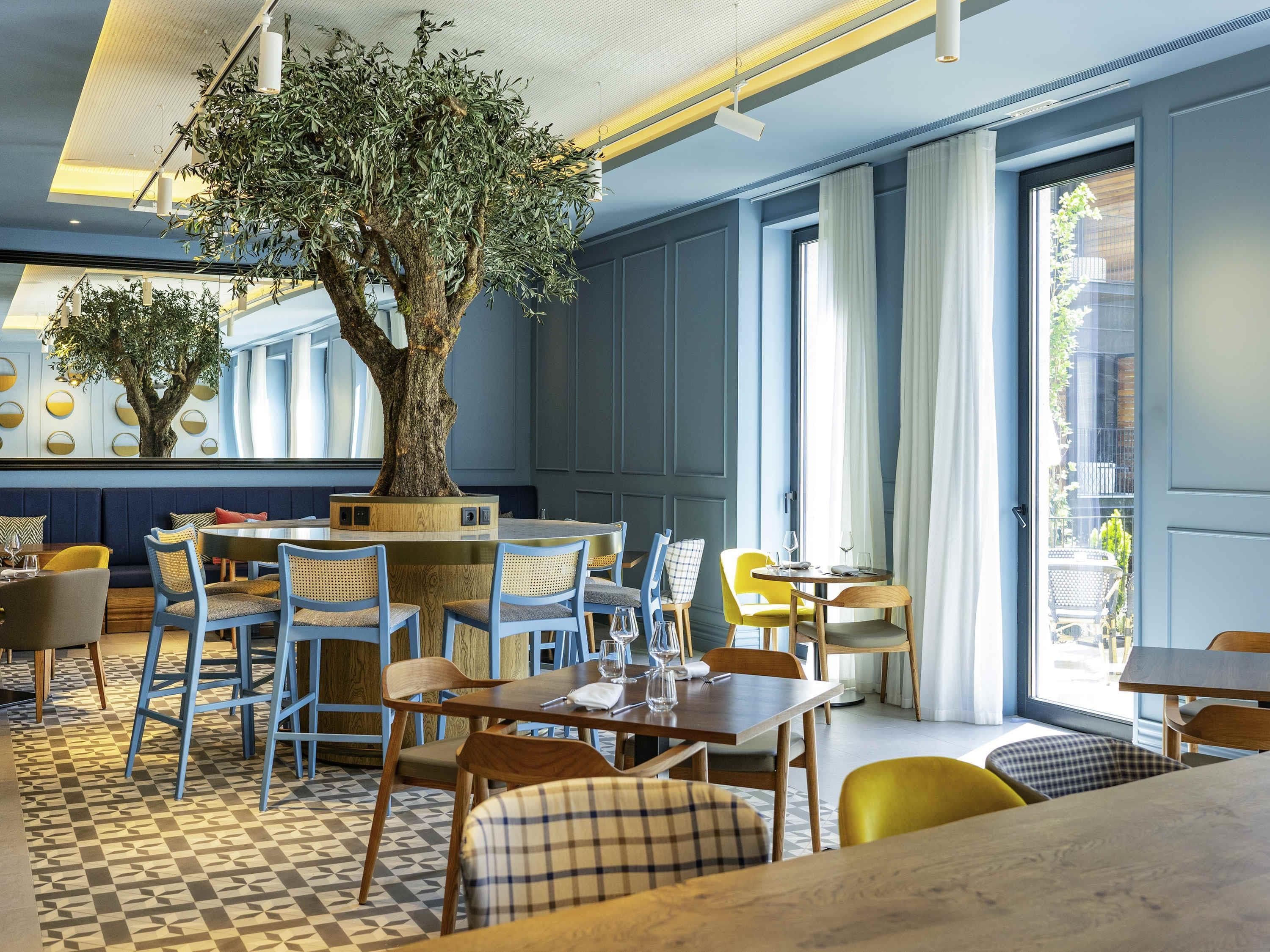 A restaurant interior with a large tree centerpiece, blue and yellow chairs, and a geometric patterned floor.