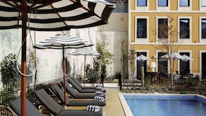 A poolside area with lounge chairs and striped umbrellas, a tiled pool, and multi-story buildings in the background.