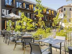 An outdoor patio with tables and chairs, surrounded by greenery and a modern building with balconies. - Mercure Porto Centro Aliados (Porto)