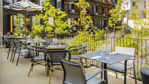 An outdoor patio with tables and chairs, surrounded by greenery and a modern building with balconies.