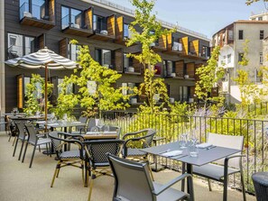 Eine Terrasse im Freien mit Tischen und Stühlen, umgeben von Grünflächen und einem modernen Gebäude mit Balkonen.