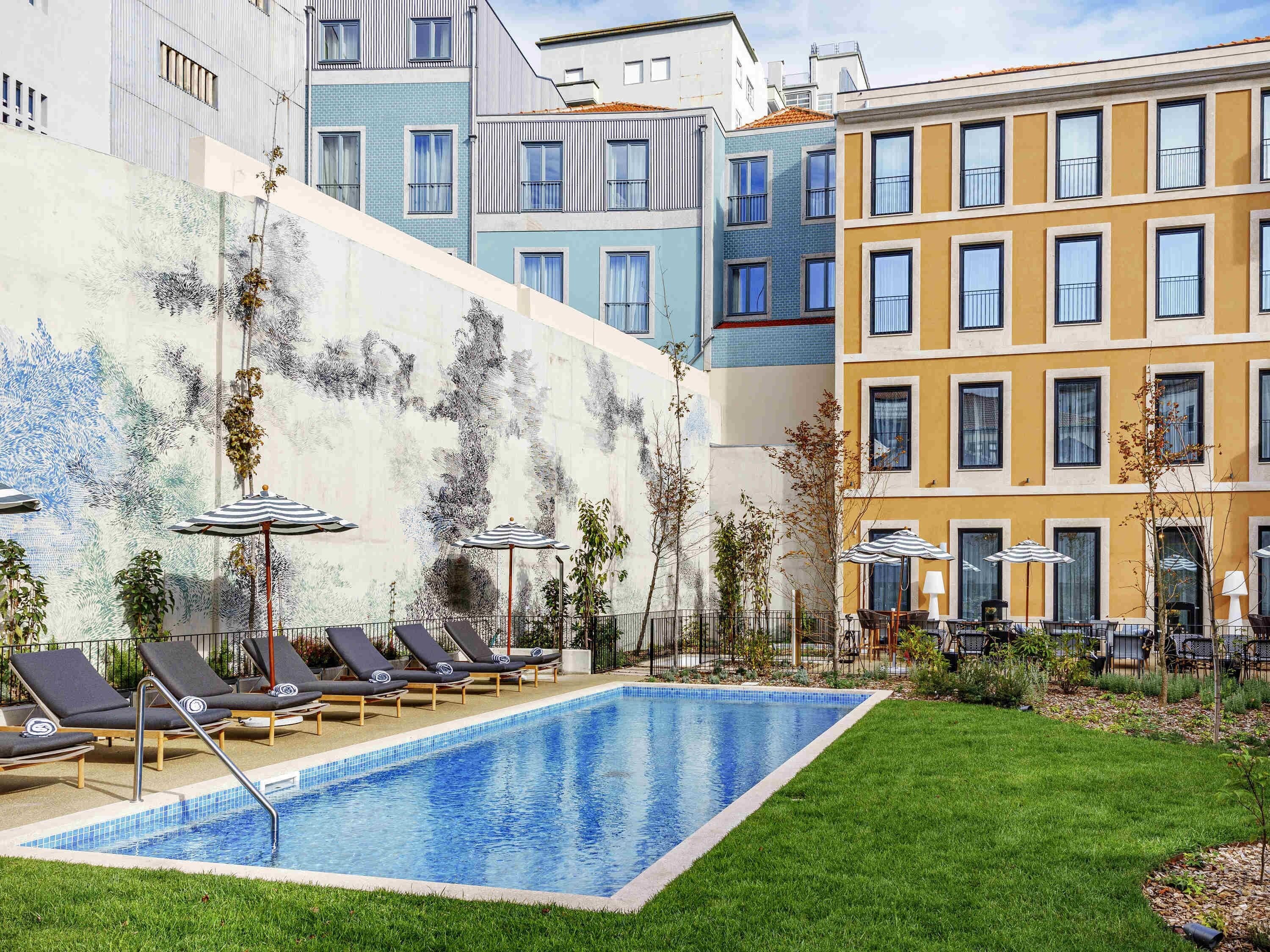 Una piscina con sillones de descanso y sombrillas, rodeada de césped y un edificio con fachadas coloridas.