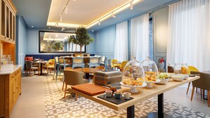 A modern hotel dining area with a buffet setup, a seating area with chairs and tables, and a large window with curtains. - Mercure Porto Centro Aliados (Porto)