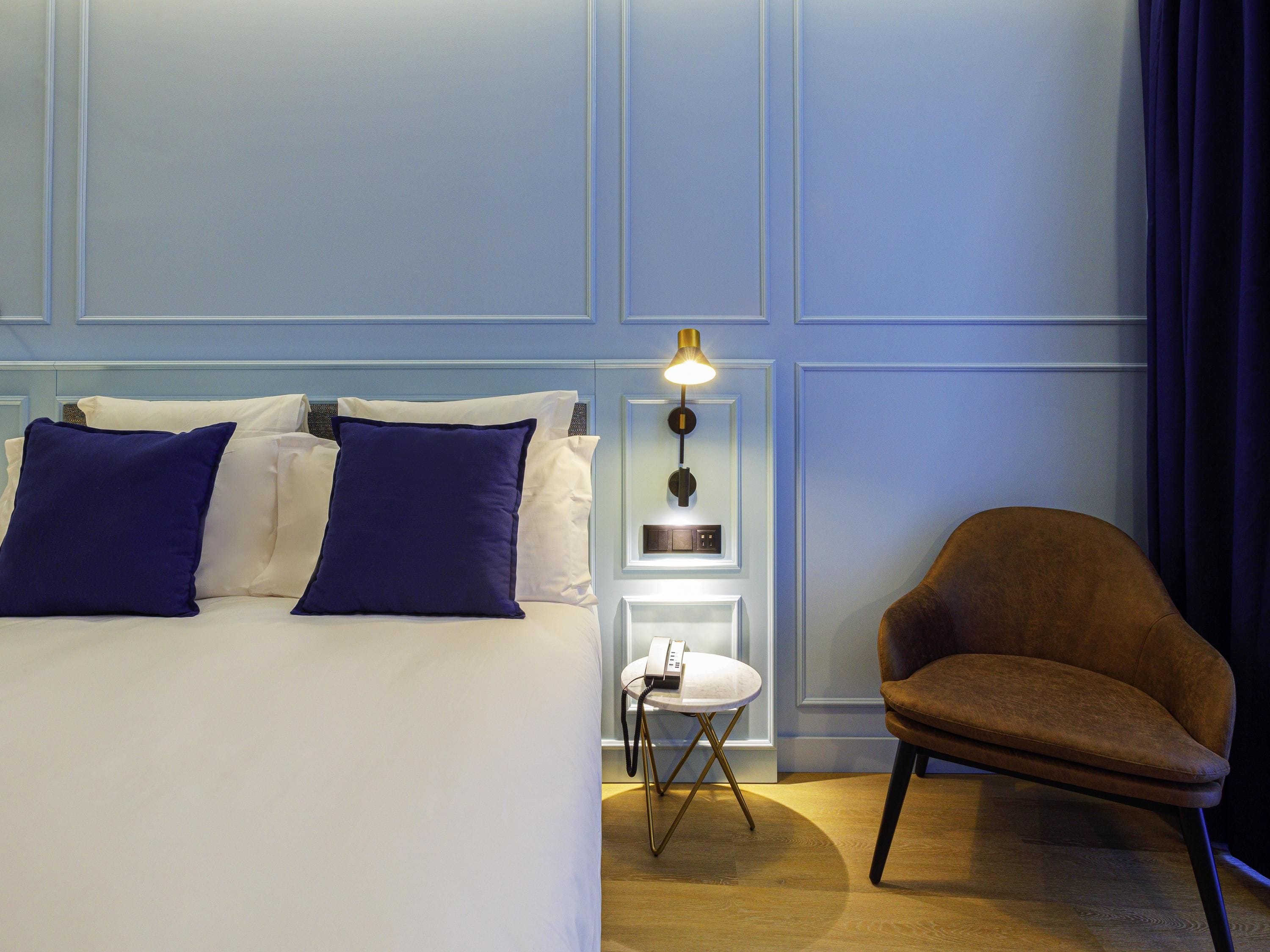 Habitación de hotel con cama, almohadas, mesita de noche, lámpara, silla y una pared con paneles.