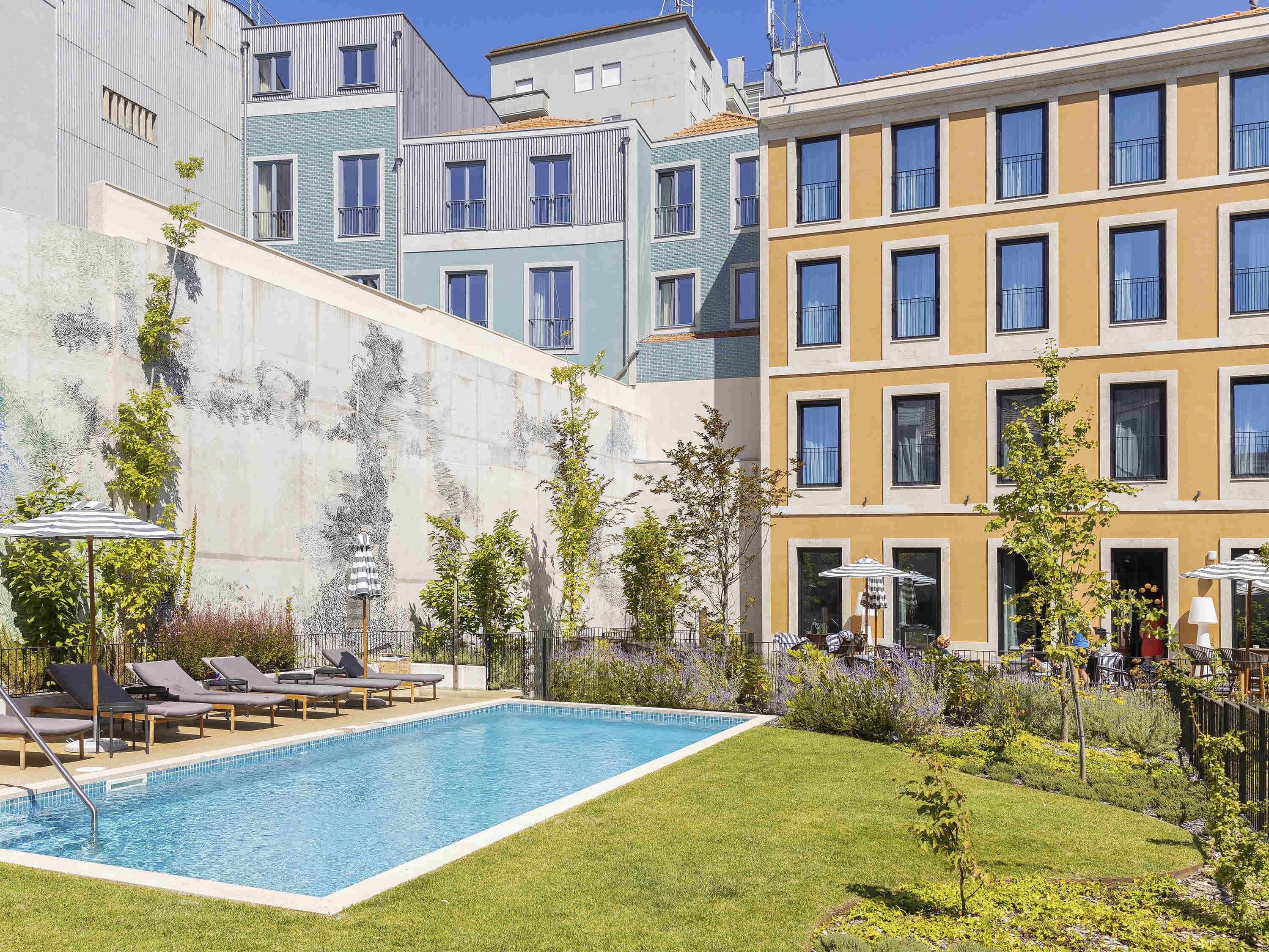 Una piscina con sillas de descanso, un jardín y edificios modernos al fondo.