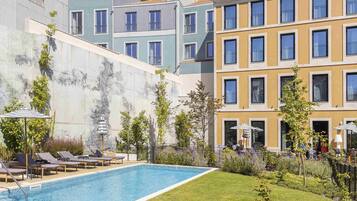 Una piscina con sillas de descanso, un jardín y edificios modernos al fondo.