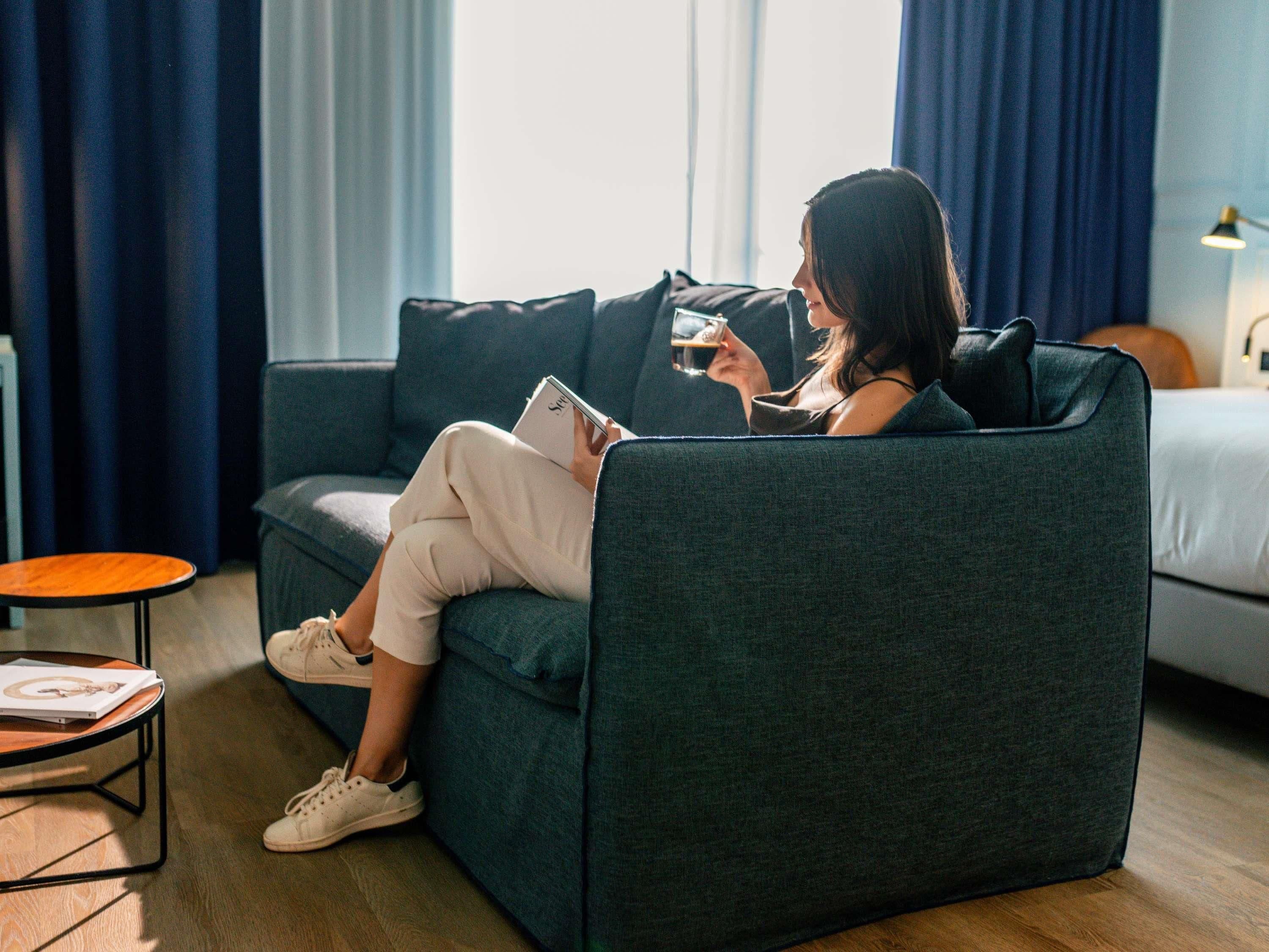 Eine Person sitzt auf einer dunkelgrünen Couch in einem Raum mit Holzboden, ein Buch und ein Getränk haltend.