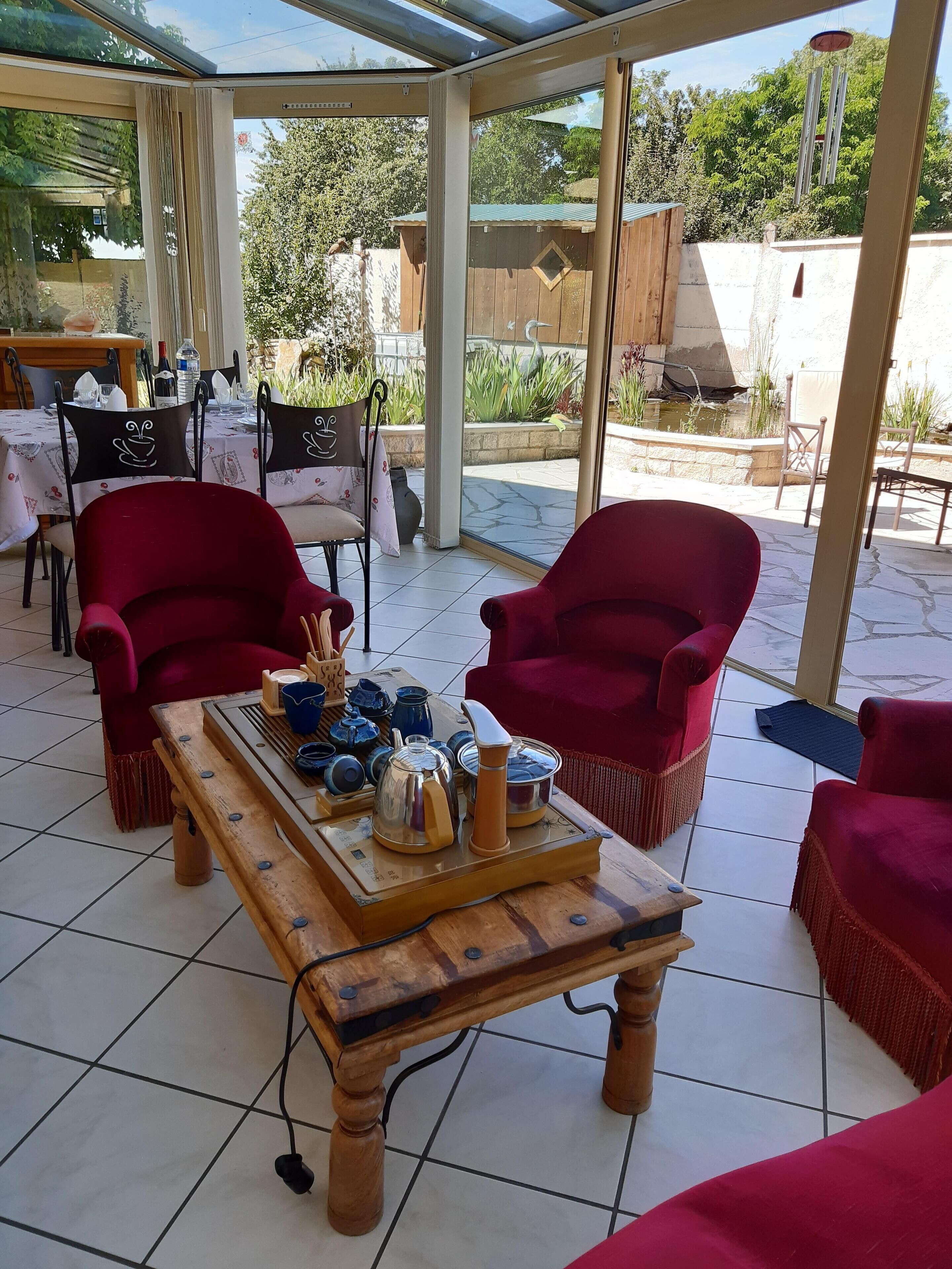 A covered outdoor seating area with red chairs, a wooden table with tea sets, and a view of a garden and trees.