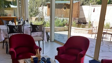 A covered outdoor seating area with red chairs, a wooden table with tea sets, and a view of a garden and trees.