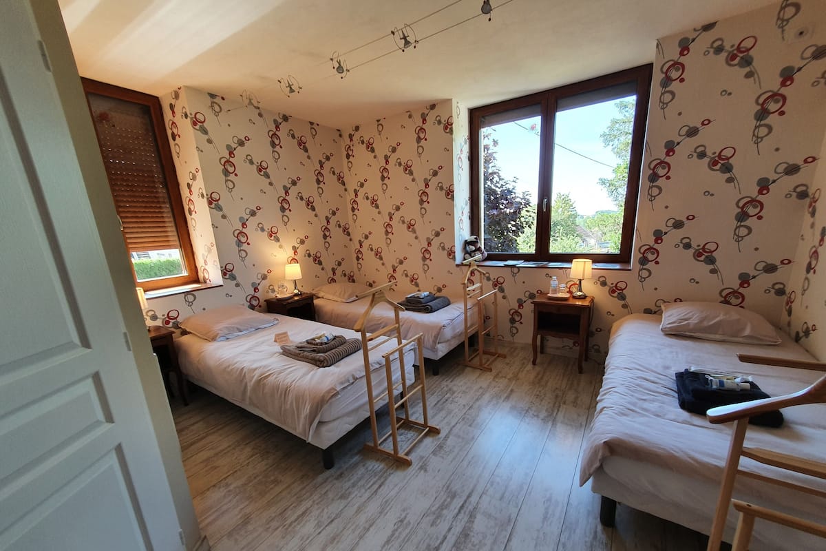 Een kamer met twee bedden, een houten trap, een klein tafeltje met een lamp en een raam met zonwering.