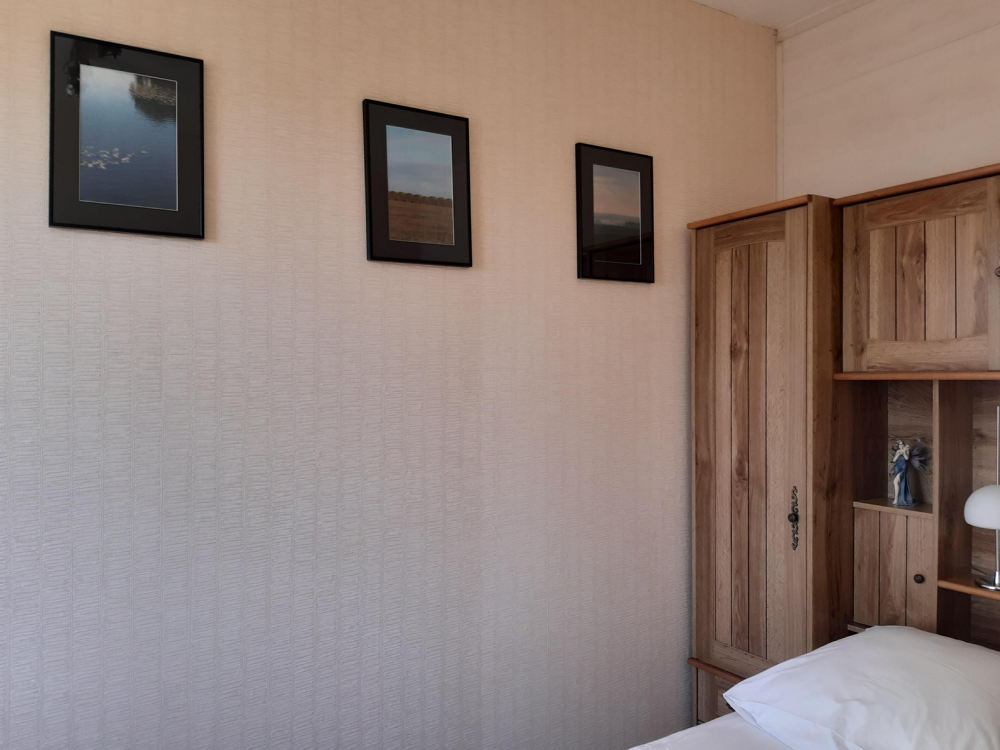 A bedroom with a bed, a wooden wardrobe, and three framed pictures on the wall.