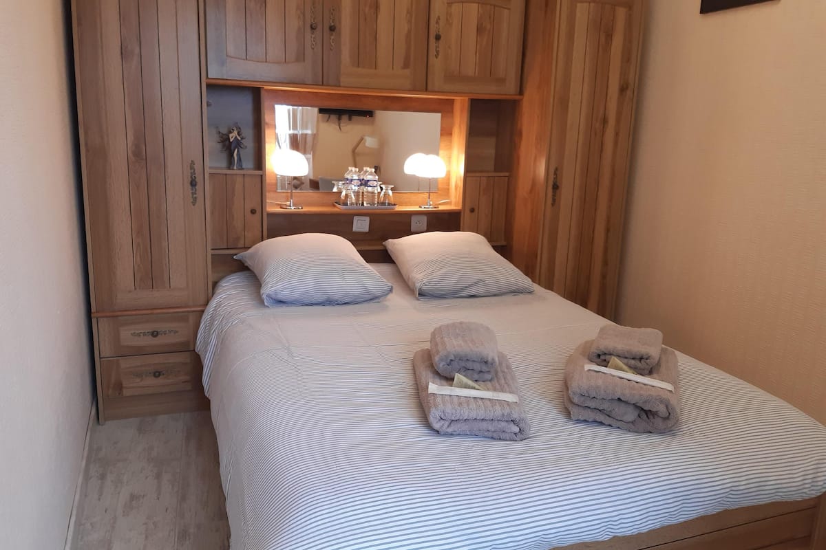 Een slaapkamer met een bed, twee kussens, een nachtkastje, een spiegel en houten kasten.