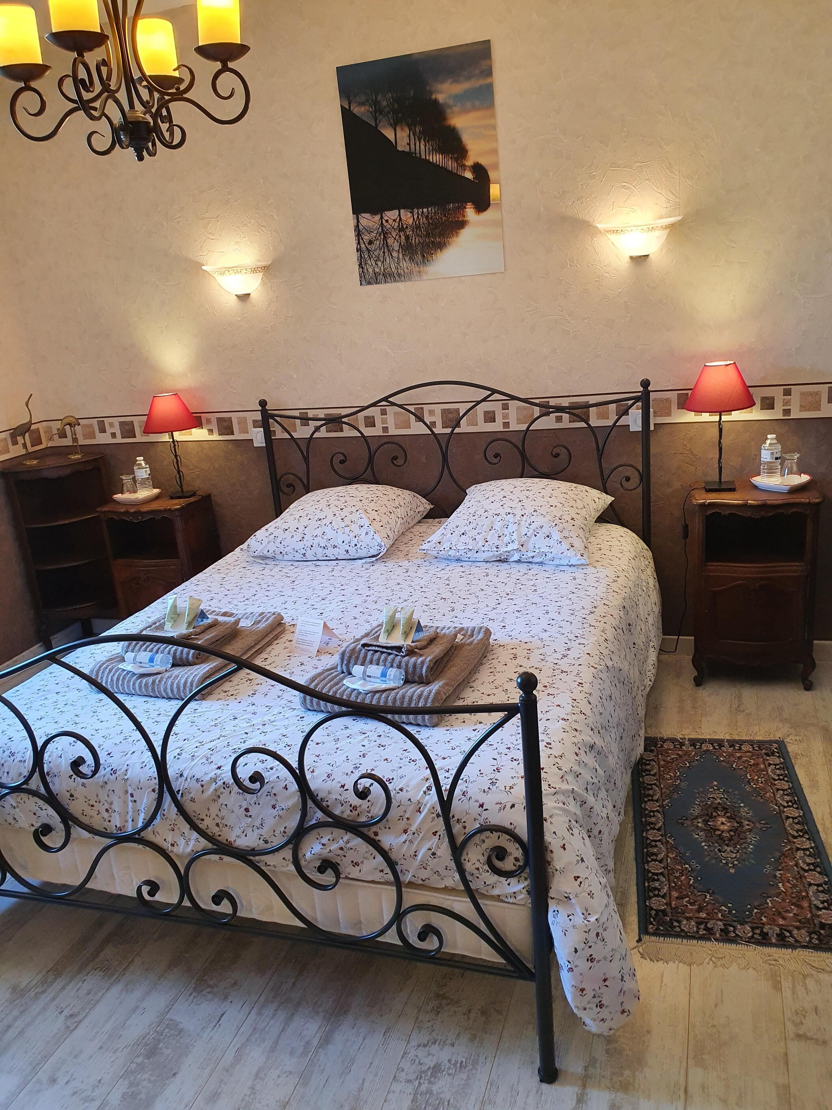 A neatly made bed with floral bedding, two bedside lamps, a nightstand with a red lamp, a wall-mounted picture, and a patterned rug.