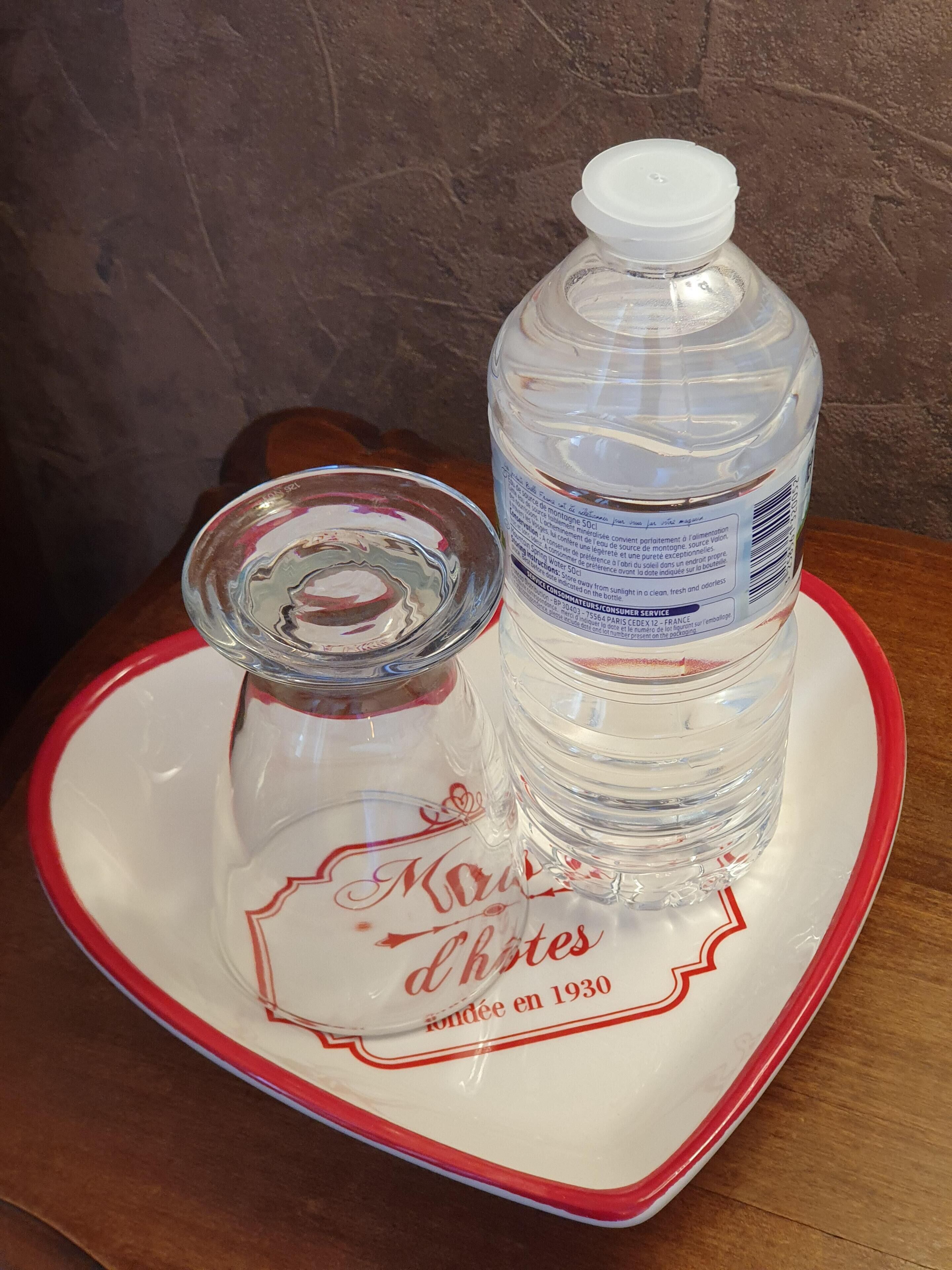 Eine klare Glasflasche mit weißem Verschluss und Glasdeckel auf einem weißen Teller mit roter Bordüre. Der Teller zeigt ein Logo und den Schriftzug 'Moulin d'Horloge fondée en 1930'.