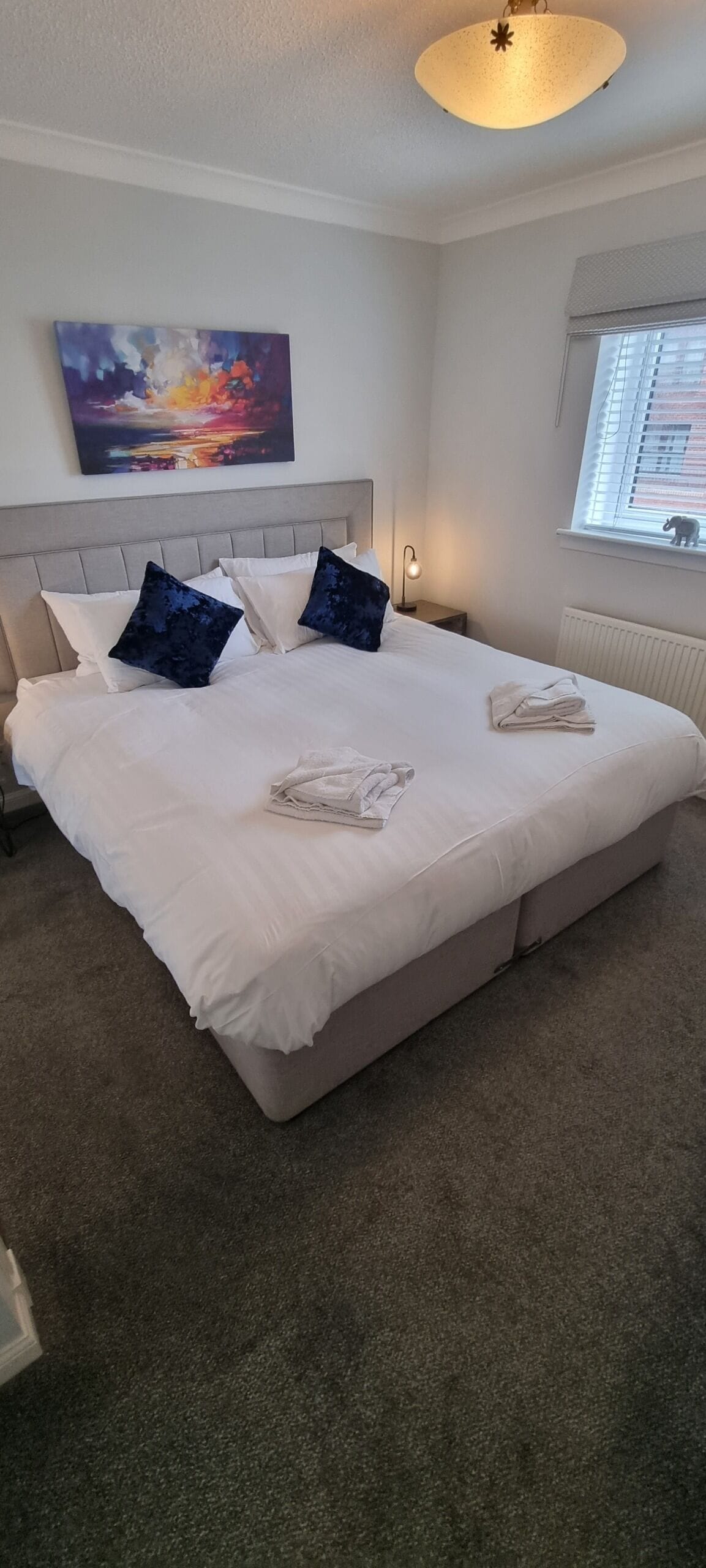 A neatly made bed with white linens and two dark blue pillows, a painting of a sunset on the wall, a bedside lamp, and a window with blinds.