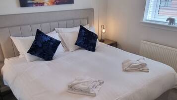 A neatly made bed with white linens and two dark blue pillows, a painting of a sunset on the wall, a bedside lamp, and a window with blinds.