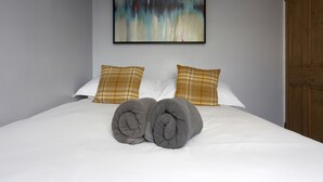 Una cama bien tendida con dos toallas enrolladas, almohadas blancas y una almohada de cuadros sobre un colchón blanco.