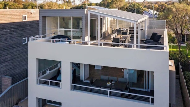 Un edificio moderno de dos pisos con balcón, ventanas grandes y mobiliario para exterior.