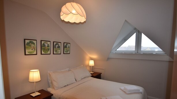 Un dormitorio con una cama, dos mesitas de noche, una lámpara, un ventanal y cuatro cuadros enmarcados en la pared.