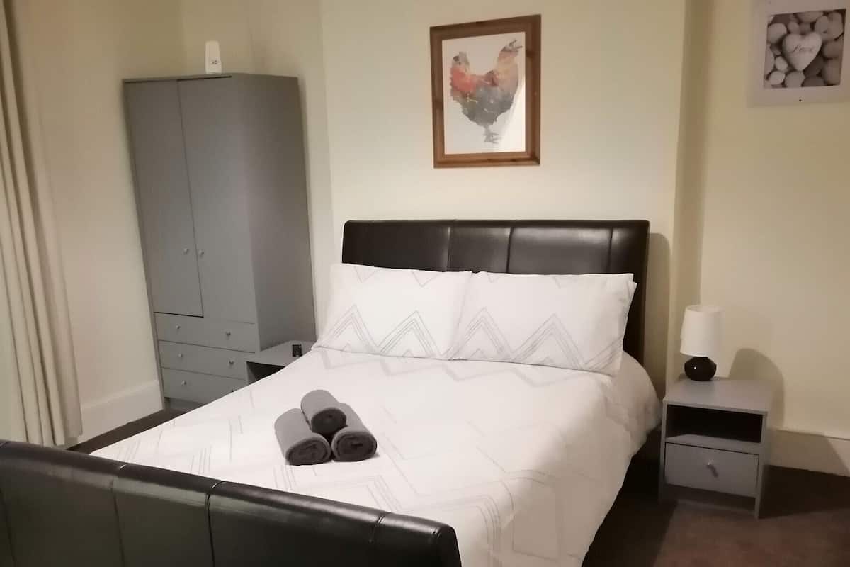 A neatly made bed with a white comforter and pillows, a grey wardrobe, a framed picture of a rooster, and a bedside table with a lamp.