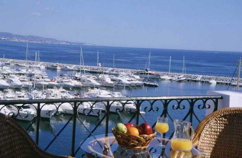 A balcony with a view of a marina, a table with a fruit bowl and drinks, and a wicker chair.