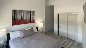 A bedroom with a bed, two bedside tables with lamps, a painting of a forest, a door, and a ceiling light fixture. - The Flatts at North Shore (Geneva)