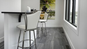 A modern bar area with a white countertop, two white bar stools, and a large window with a view of the street. - The Flatts at North Shore (Geneva)