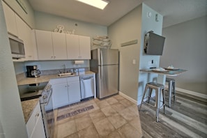 A compact kitchen with white cabinets, a stainless steel refrigerator, a coffee maker, and a small dining area with stools.