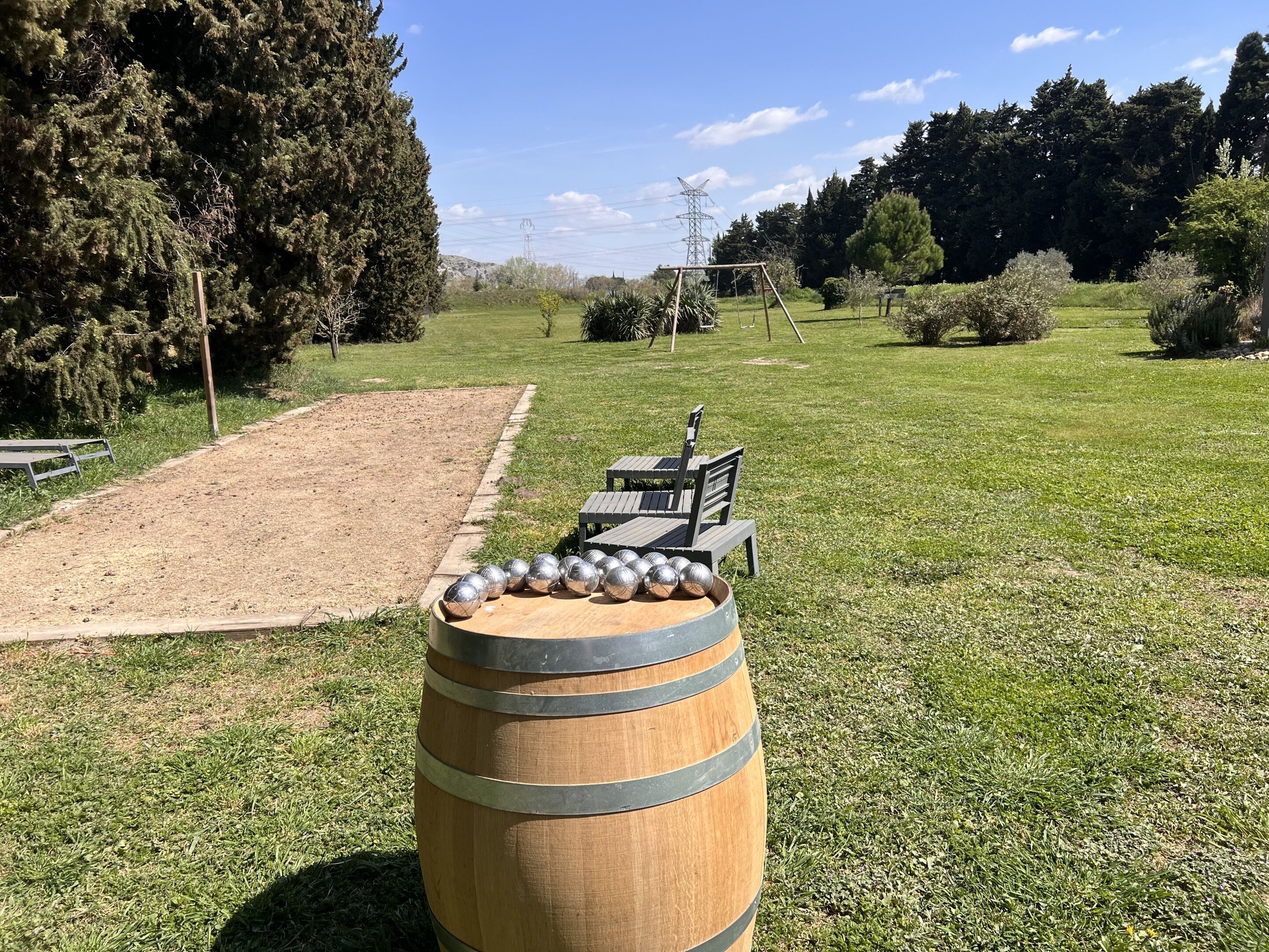 Un tonneau en bois cerclé de métal, un banc et une balançoire dans un champ herbeux, avec des arbres et une ligne électrique en arrière-plan.