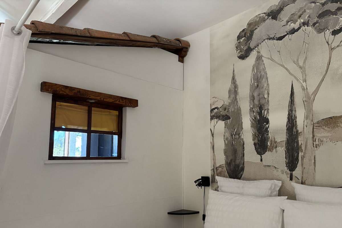 A bedroom with a bed, white pillows, a window with a wooden frame, and a wall with a tree mural.