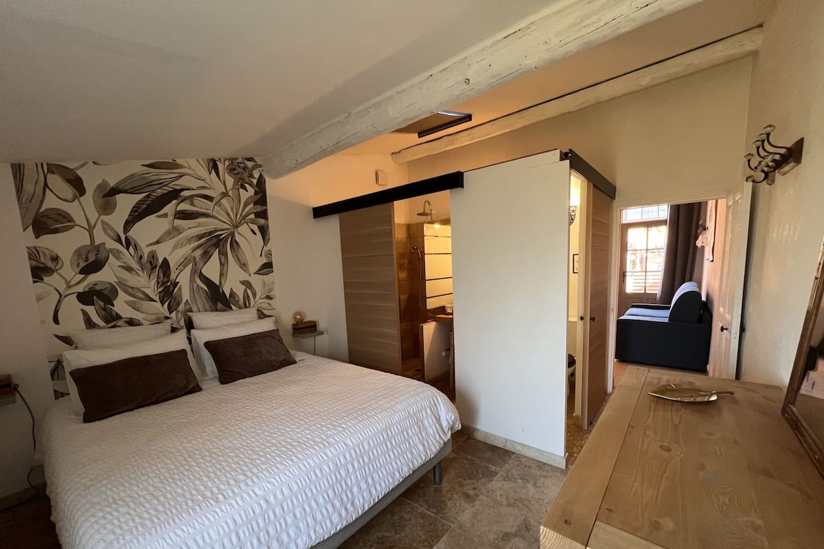 A bedroom with a bed, a wooden dresser, a bathroom with a shower, and a view of the outside.