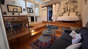 A cozy living room with a dark sofa, a round coffee table, and a patterned rug. A kitchen with a dining area is visible in the background. - Mas D'auseu (Plan-d'Orgon)