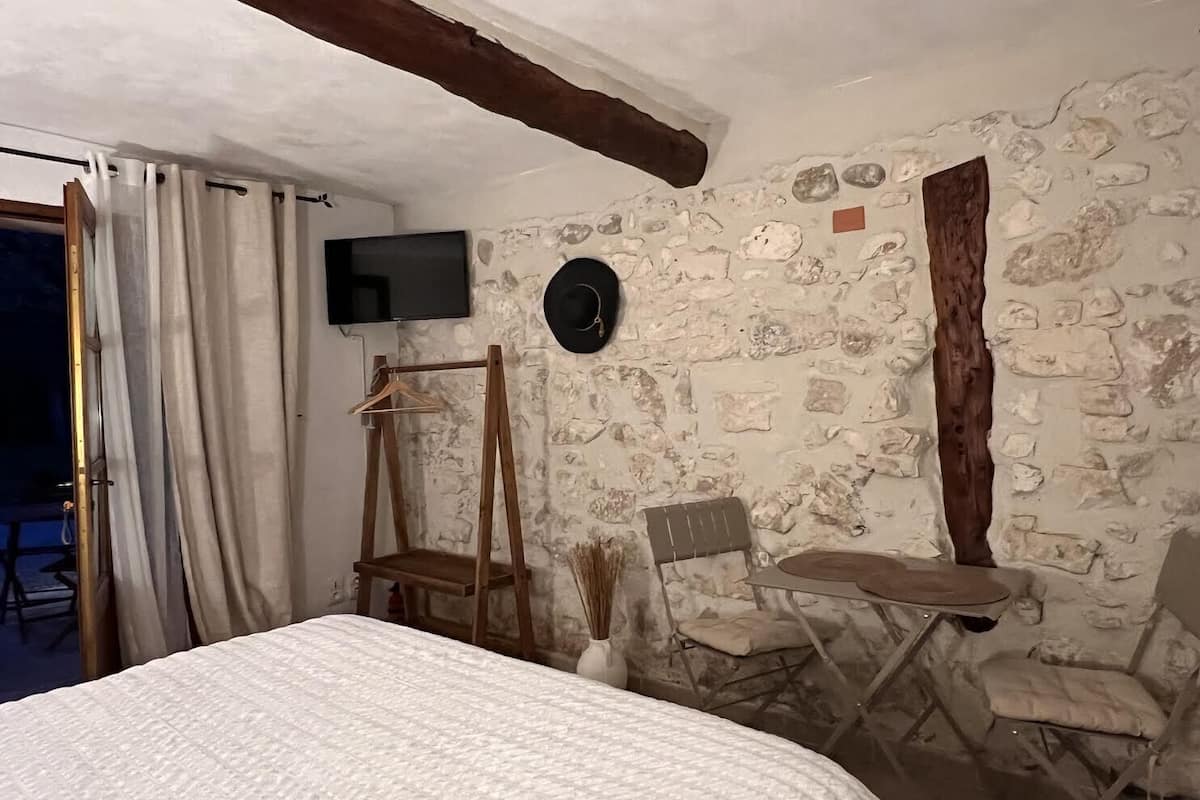 A bedroom with a bed, a TV mounted on the wall, a wooden chair, and a small table.