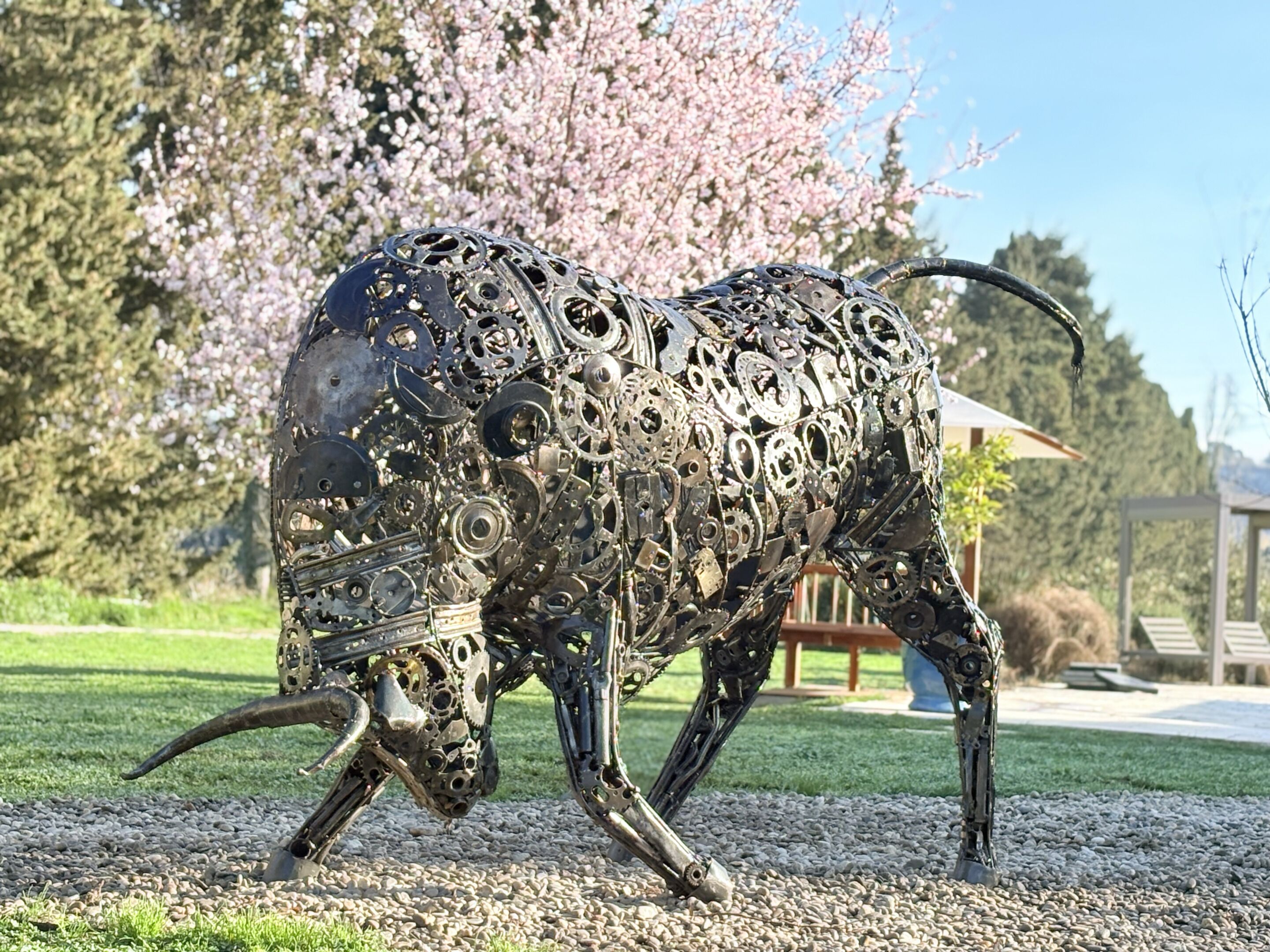 Una escultura de toro hecha con engranajes y cadenas de metal en un entorno exterior, con una zona de césped y árboles al fondo.